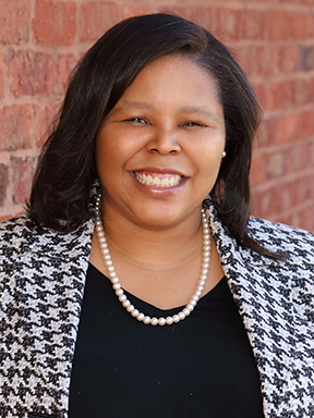 A young woman in a houndstooth jacket in front of a brick wall.