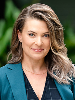 A woman with long, flowing hair wearing a teal blazer with a black patterned blouse stands in front of greenery.