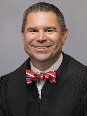 A man wearing a black judge's robe and a red striped bow tie smiling warmly.