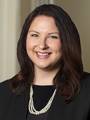 A woman with long brown hair in a black business suit wearing pearls and smiling warmly at the camera.