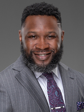 A bearded man in a gray striped suit and purple paisley necktie.