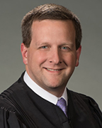 A man in a black judge's robe wearing a white shirt and purple tie.